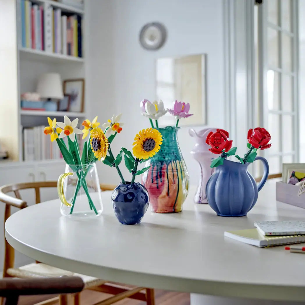 Various LEGO Botanicals models, including sunflowers and roses, displayed in different vases on a white table.