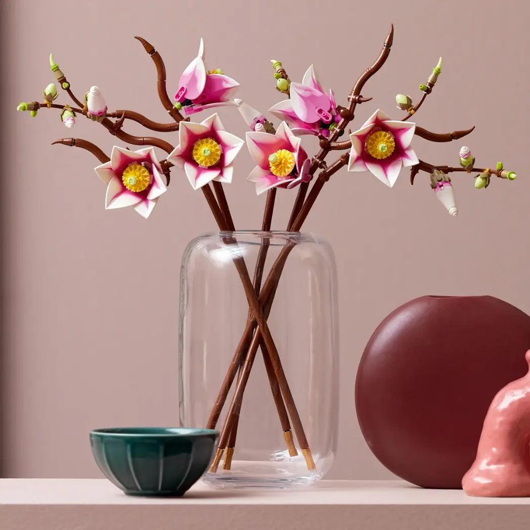 LEGO Magnolia Branches in a clear glass vase on a shelf next to a green bowl and a round reddish-brown vase.
