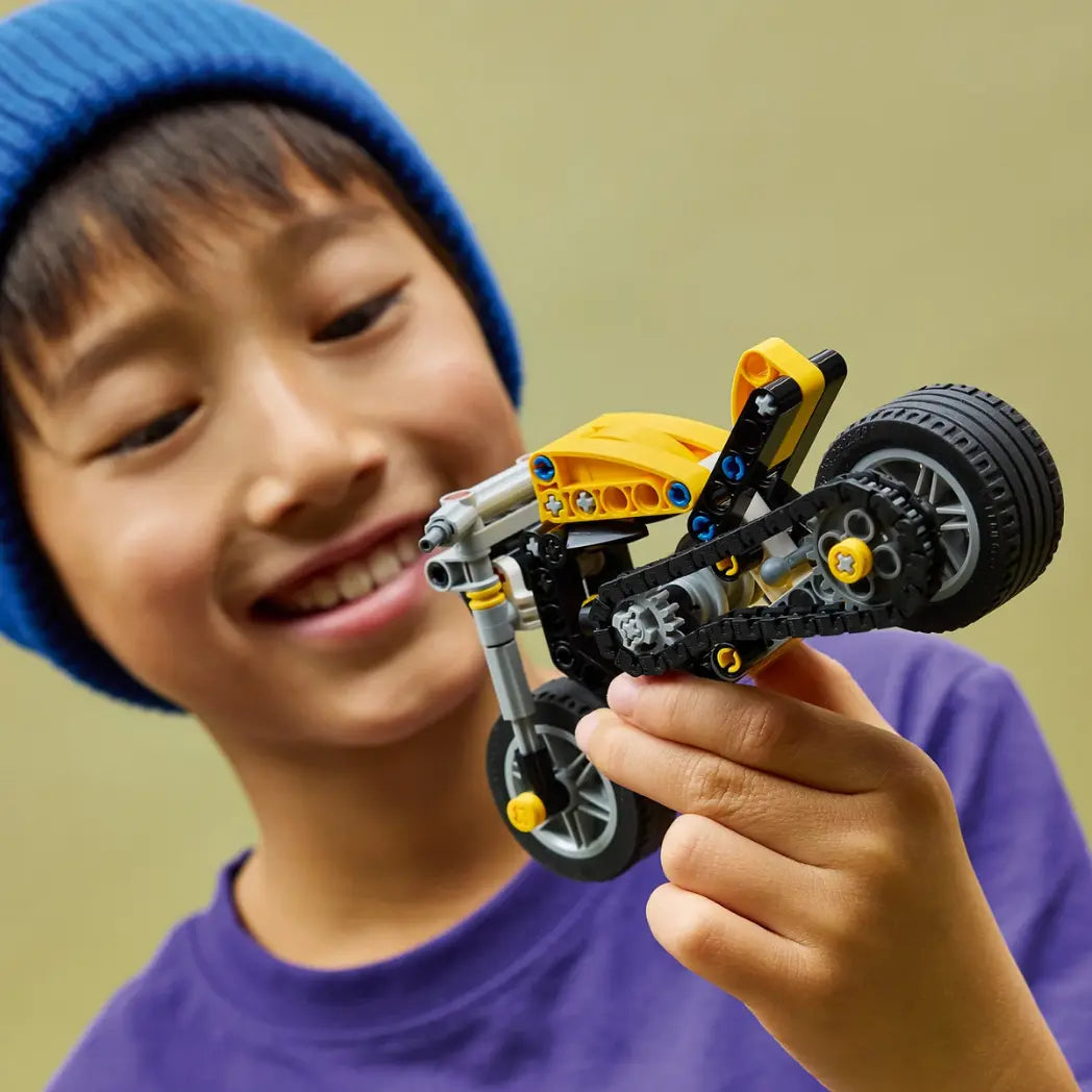A young boy wearing a blue beanie smiles while holding a built LEGO Technic yellow motorcycle.